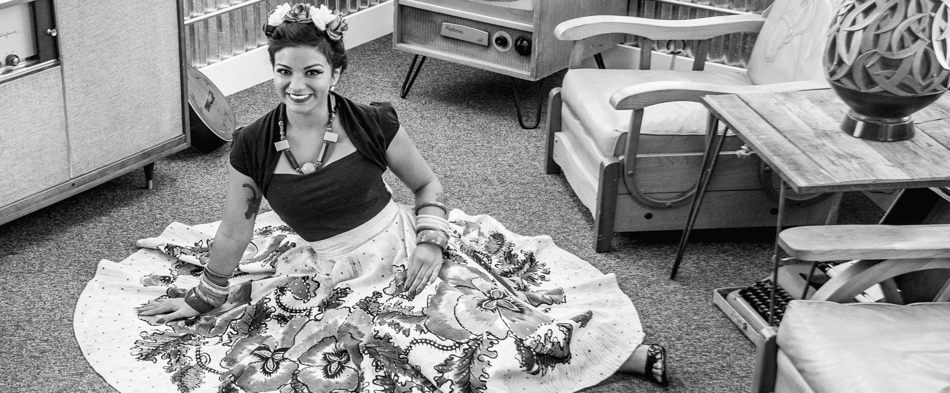 A woman in a floral dress sits on the floor, smiling amidst vintage furniture in a black-and-white setting.