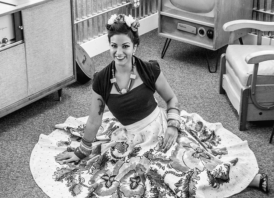 A woman in traditional attire sits on the floor, smiling against a retro backdrop.
