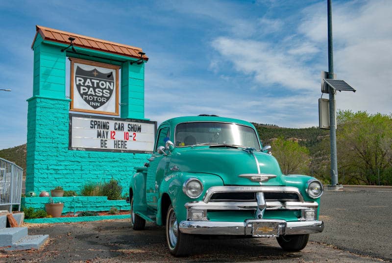 A vintage turquoise truck parked in front of the Raton Pass Motor Inn, advertising a spring car show.