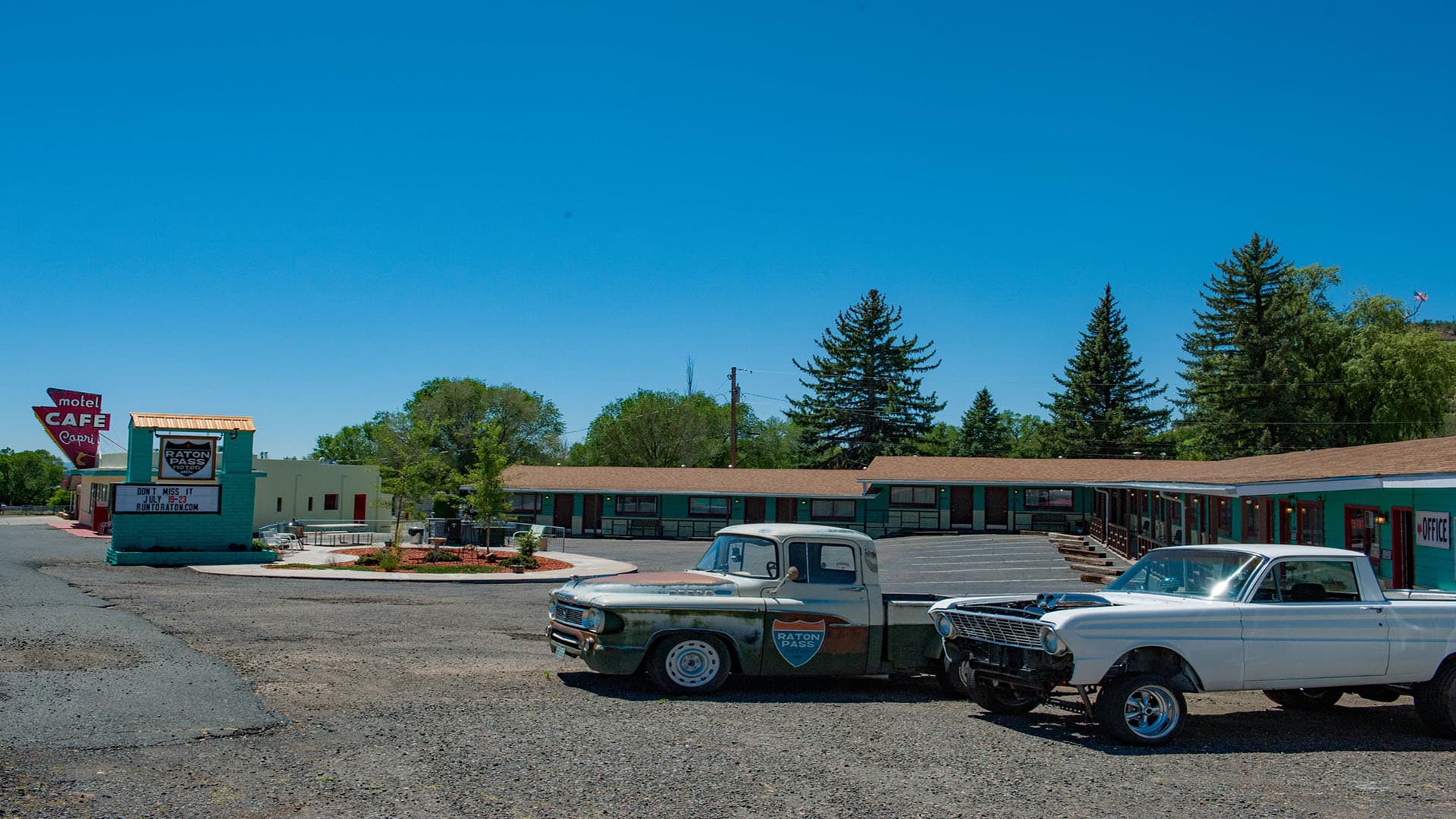 A vintage pickup and a classic car parked in front of a retro motel under a clear blue sky.