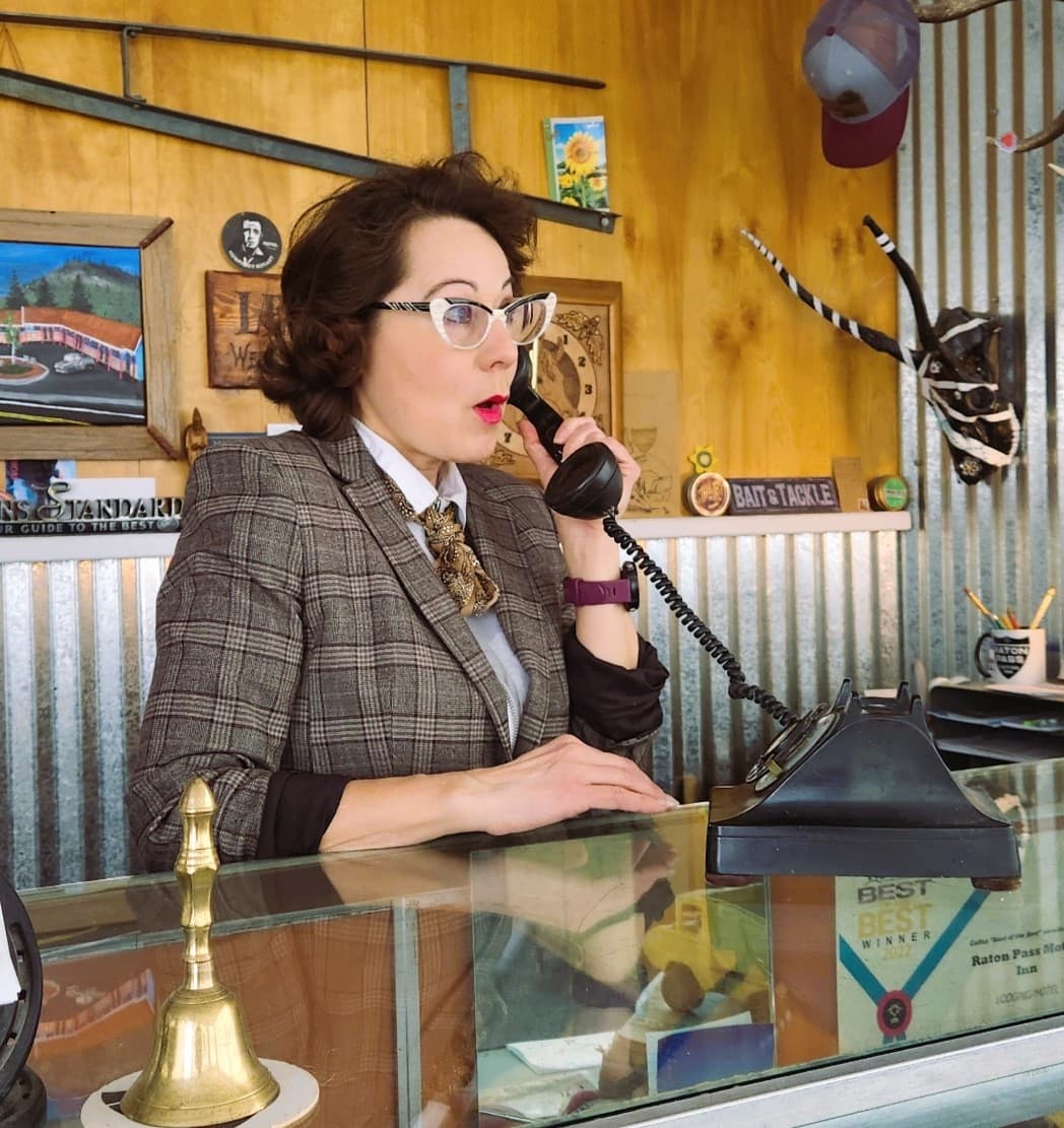 A woman in a plaid suit speaks into a vintage rotary phone at a reception desk. A woman in a plaid suit speaks into a vintage rotary phone at a reception desk.