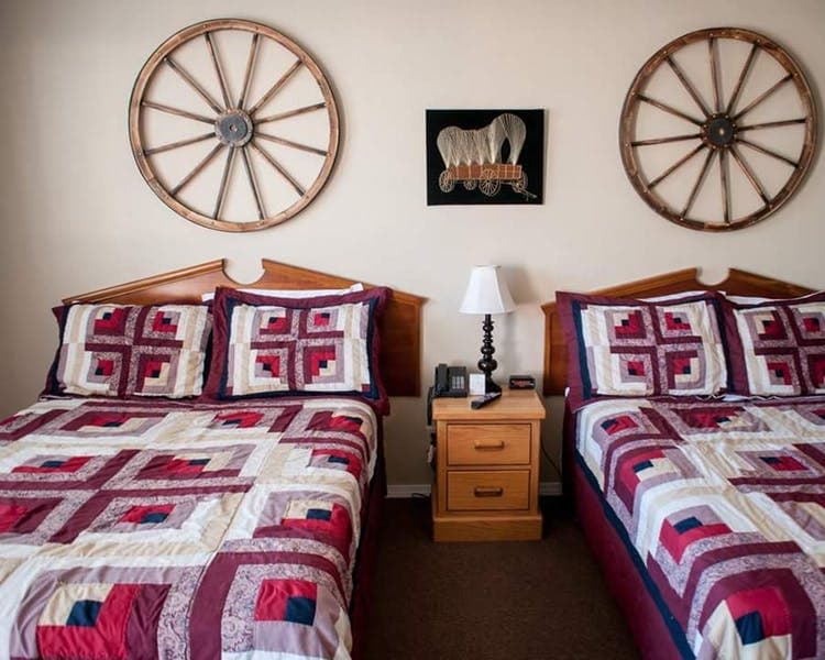 A cozy bedroom featuring two beds with patterned quilts, a bedside table with a lamp, and decorative wagon wheel wall art.